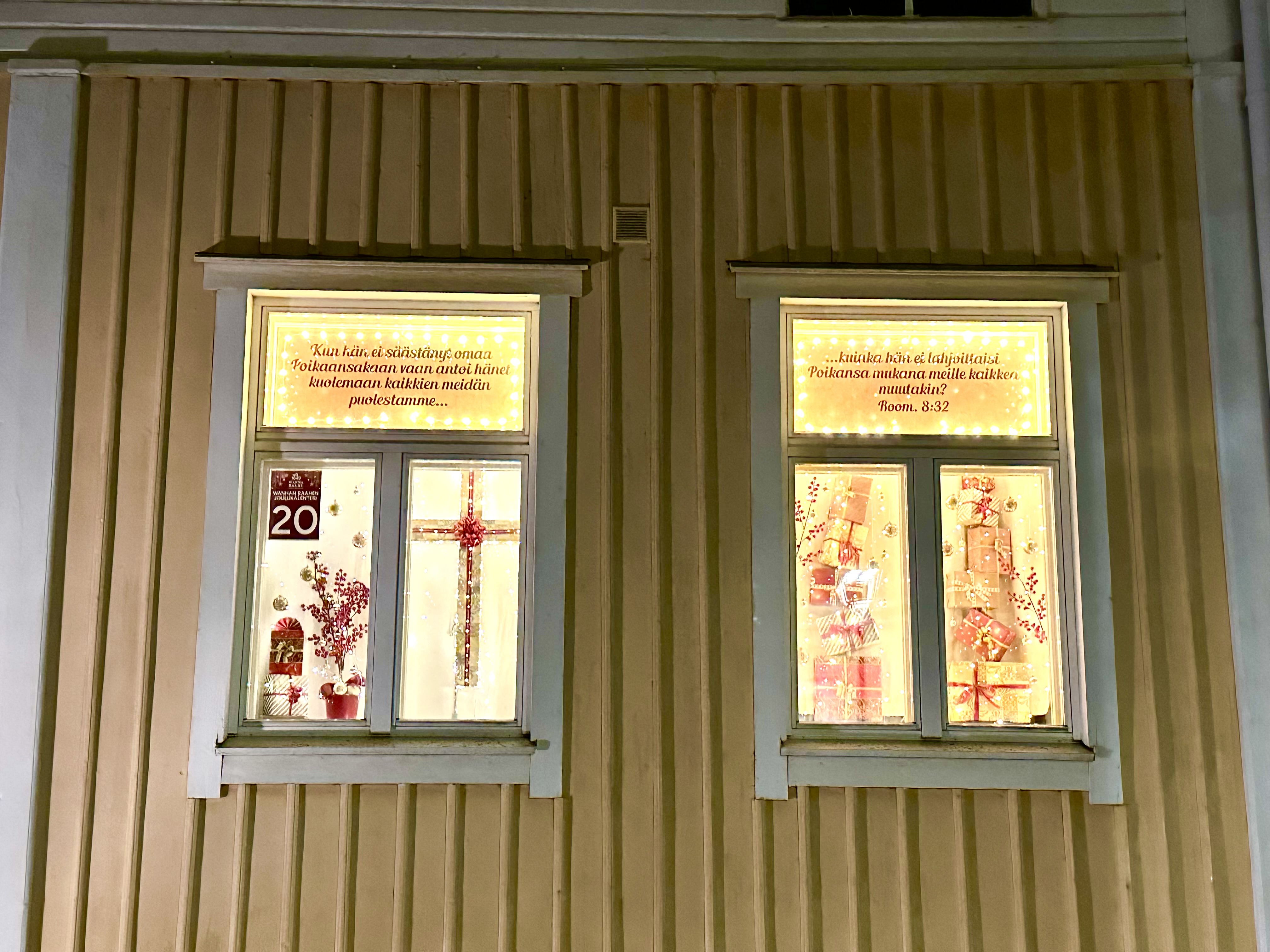 Luukku 20. Raamatun teksti ikkunassa, jouluinen risti sekä lahjapaketteja ja lumihiutaleita. 
